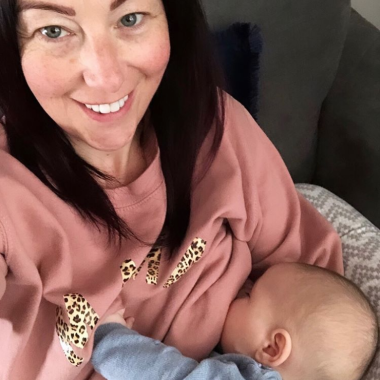 A woman taking a selfie from a high angle, she is wearing a pink sweater and breastfeeding a baby