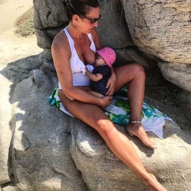 A woman in a white swimsuit, sat in the shade of some large rocks on a beach, breastfeeding a baby