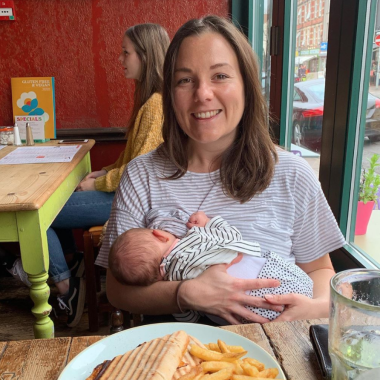 A woman sat in a cafe with a plate of chips and a sandwich in front of her, breastfeeding a baby