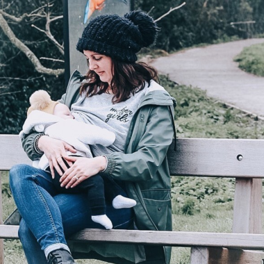 A woman sat on a bench outdoors, lifting her sweater to breastfeed a baby in a jacket and leggings 